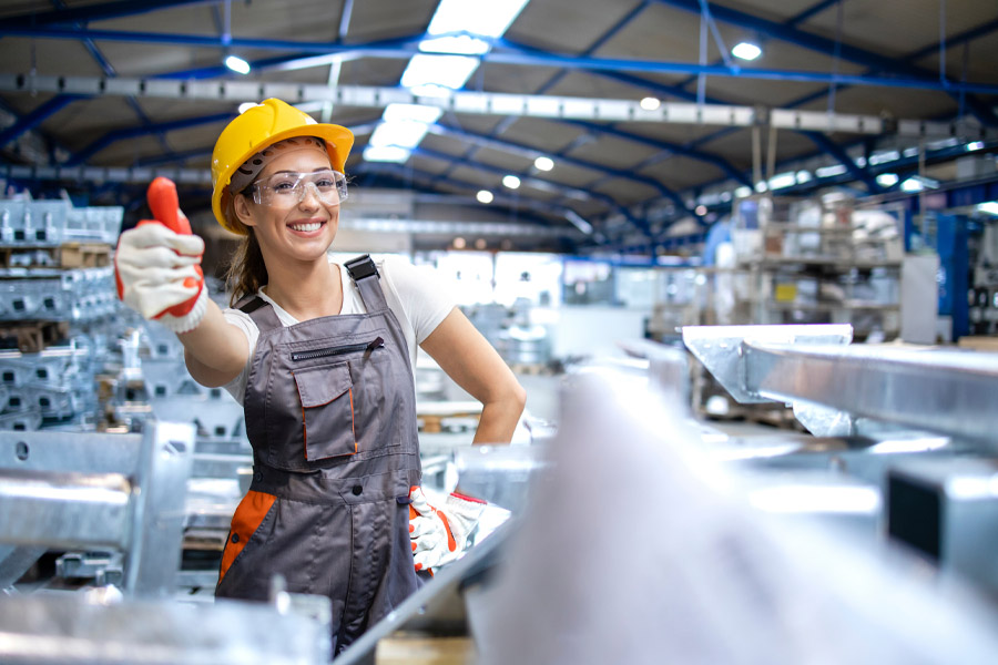 Eine Frau mit Bauhelm und Schutzbrille streckt den Daumen nach oben, während sie zwischen Metallträgern in einer Fabrik steht. Sie trägt einen grauen Arbeitsoverall und Handschuhe und lächelt selbstbewusst vor der verschwommenen industriellen Kulisse, was die Effizienz der modernen Beleuchtung Industriehallen unterstreicht.