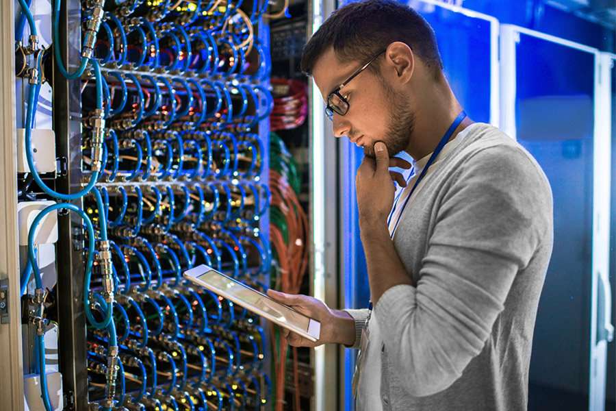 Ein Mann mit Brille steht in einem Serverraum, hält ein Tablet in der Hand und inspiziert aufmerksam Netzwerkkabel und Geräte. Das blaue Licht unterstreicht die Bedeutung der Sicherheitsbeleuchtung in Unternehmen für einen sicheren Arbeitsplatz.