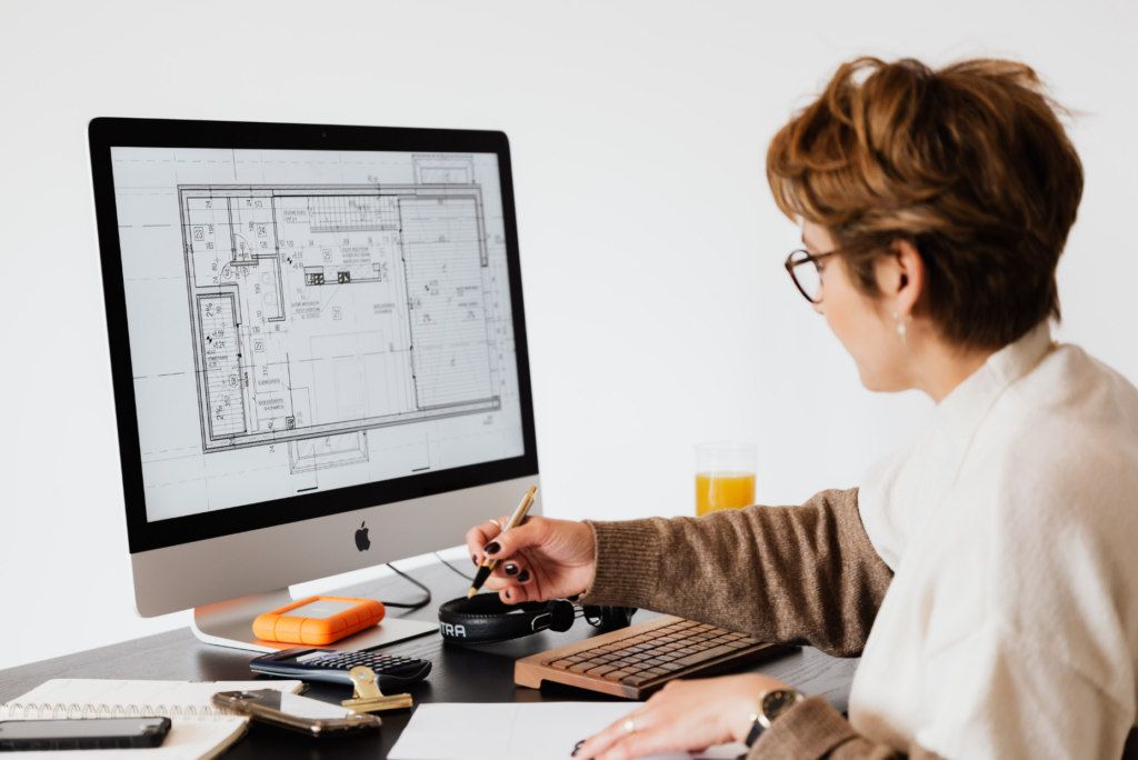 Eine Person mit kurzen Haaren und Brille arbeitet an einem Schreibtisch, sieht sich architektonische Entwürfe und Lichtplanungen an, die auf einem großen Computermonitor angezeigt werden, macht sich Notizen und hat Büromaterial und ein Glas Orangensaft dabei.