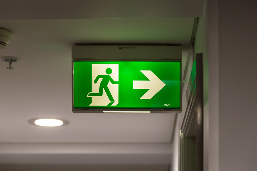 Ein grün beleuchtetes Notausgangsschild mit einer laufenden Figur und einem nach rechts zeigenden Pfeil, das an der Decke eines Flurs angebracht ist, dient als wichtige Sicherheitsbeleuchtung in Unternehmen und weist auf den nächsten Ausgang hin.