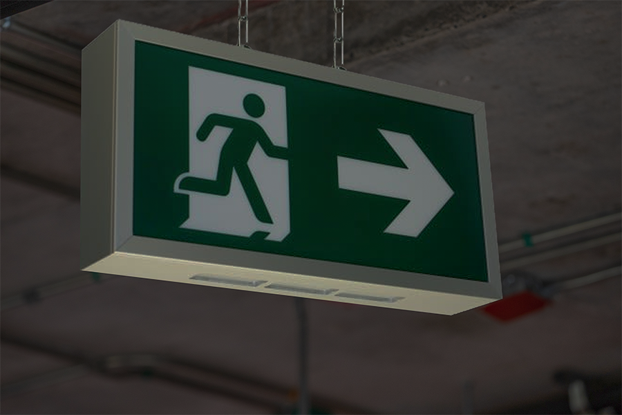 An einer Decke mit freiliegenden Rohren hängt ein grünes Notausgangsschild, das eine laufende Figur zeigt, die sich durch eine Tür bewegt, mit einem großen weißen Pfeil, der nach rechts zeigt.