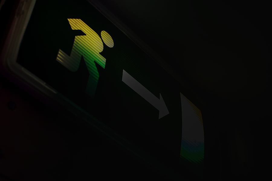 Ein leuchtend grünes Notausgangsschild zeigt eine laufende Figur und einen nach rechts gerichteten Pfeil, der die Richtung zum Ausgang angibt. Der Hintergrund ist dunkel, wodurch sich das Schild deutlich abhebt.