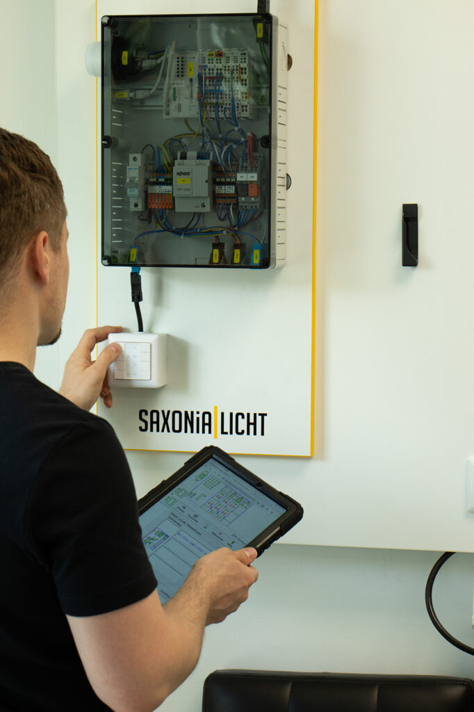 Eine Person inspiziert mithilfe eines Tablets einen offenen Schaltschrank an der Wand über einem Schild mit der Aufschrift „SAXONIA LICHT“. Im Inneren des Schaltschranks sind verschiedene Kabel und Komponenten sichtbar.