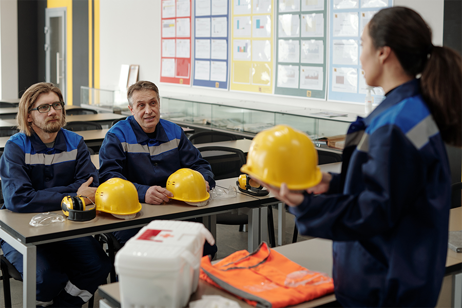 Drei Personen in blauen Uniformen befinden sich in einem Klassenzimmer. Zwei von ihnen sitzen an Tischen mit gelben Schutzhelmen und Sicherheitsausrüstung und hören einer stehenden Frau zu, die einen Schutzhelm trägt. An der Wand hinter ihnen sind Sicherheitsplakate zu sehen.
