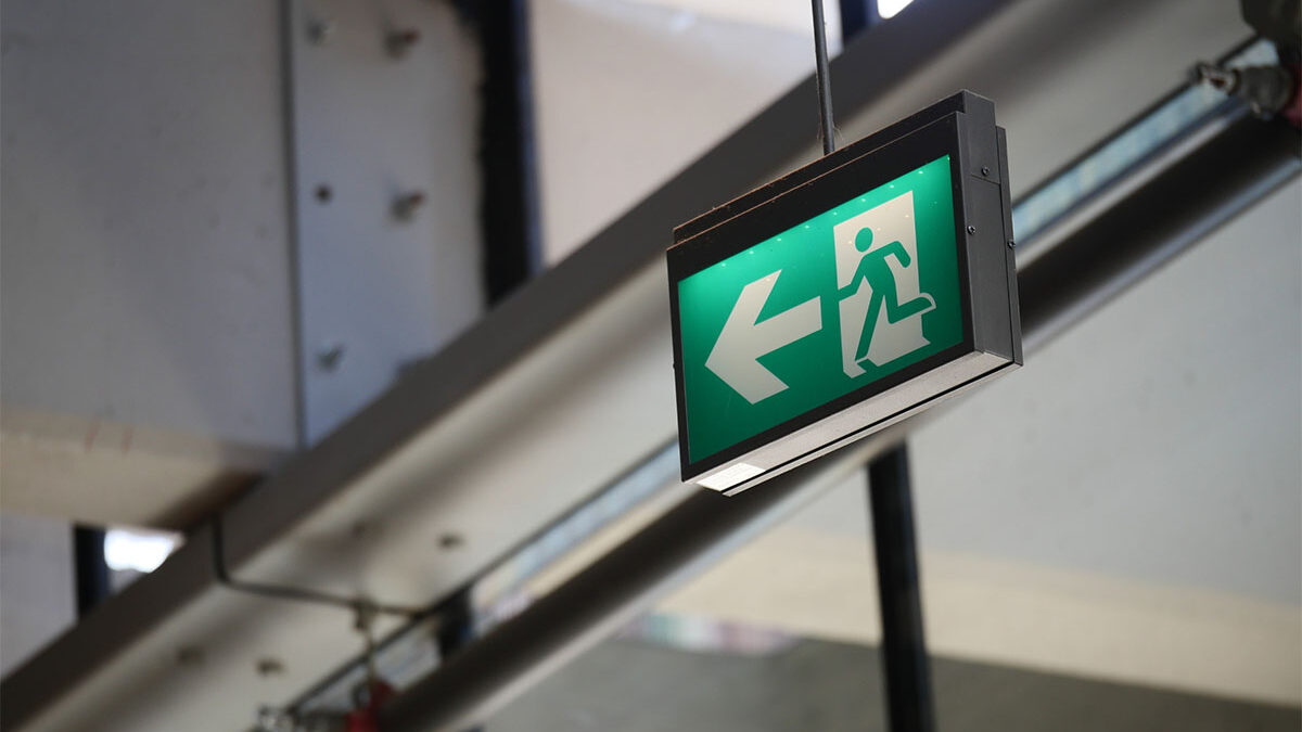 Ein grünes Notausgangsschild mit einer laufenden Figur und einem Pfeil nach links hängt von der Decke in der Nähe großer Fenster und freiliegender Rohre.