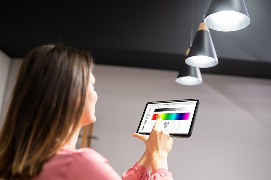 Eine Frau verwendet ein Tablet, um die intelligente Beleuchtung anzupassen, indem sie die Farb- und Helligkeitseinstellungen auf dem Bildschirm ändert, während sie unter modernen Hängelampen steht.
