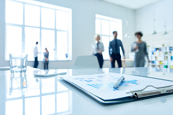 Ein Klemmbrett mit Tabellen und einem Stift liegt auf einem glänzenden Schreibtisch in einem hellen Büro. Im Hintergrund stehen vier Personen und unterhalten sich an großen Fenstern, die leicht unscharf sind.