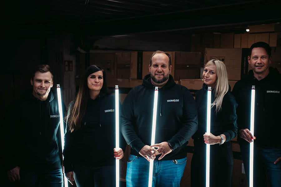 Fünf Menschen in schwarzen Kapuzenpullovern stehen zusammen und halten vertikale LED-Lichtröhren, die ihre Gesichter mit einem dramatischen Schein beleuchten, der an die Beleuchtung in riesigen Industriehallen erinnert, und das alles vor einem Hintergrund aus gestapelten Pappkartons und einer düsteren Umgebung.