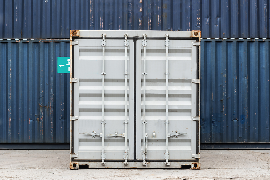 Ein geschlossener grauer Schiffscontainer steht auf einer Betonfläche, vor einem größeren, dunkelblauen Schiffscontainer, der dahinter gestapelt ist. Links ist teilweise ein kleines türkisfarbenes Schild mit einem weißen Pfeil zu sehen.