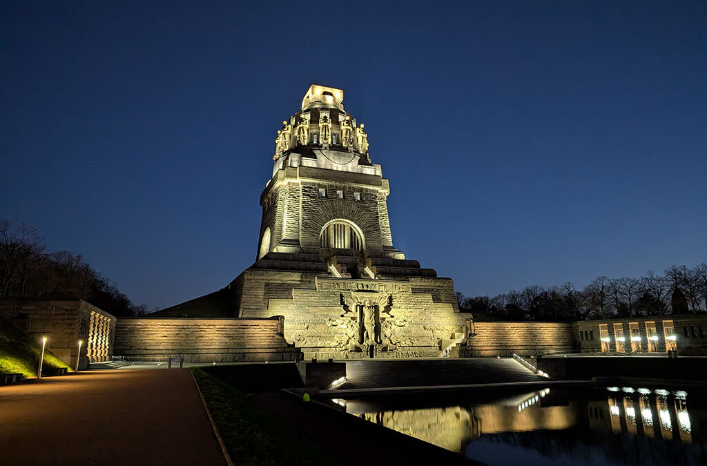 Ein großes, nachts beleuchtetes Steinmonument mit kunstvollen Schnitzereien und Statuen an seinem Sockel, das sich in einem stillen Wasserbecken vor einem dunkelblauen Himmel spiegelt.