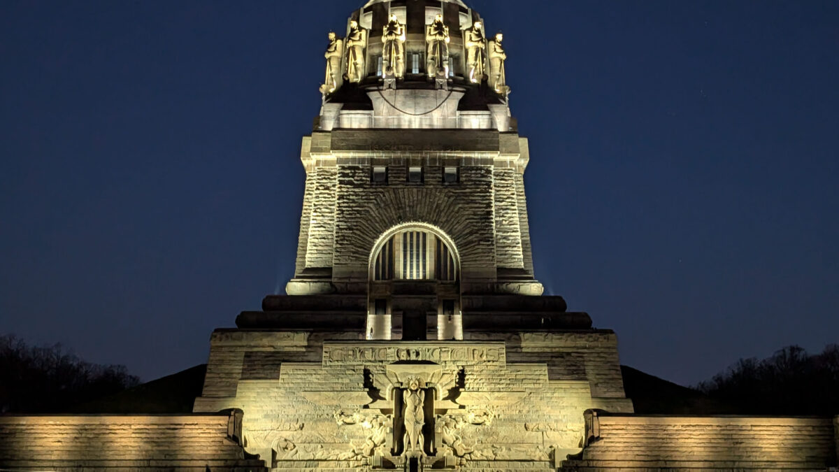 Das beleuchtete Völkerschlachtdenkmal in Leipzig, Deutschland, erhebt sich bei Nacht, und an seiner massiven Steinfassade sind Statuen und detaillierte Schnitzereien zu sehen.