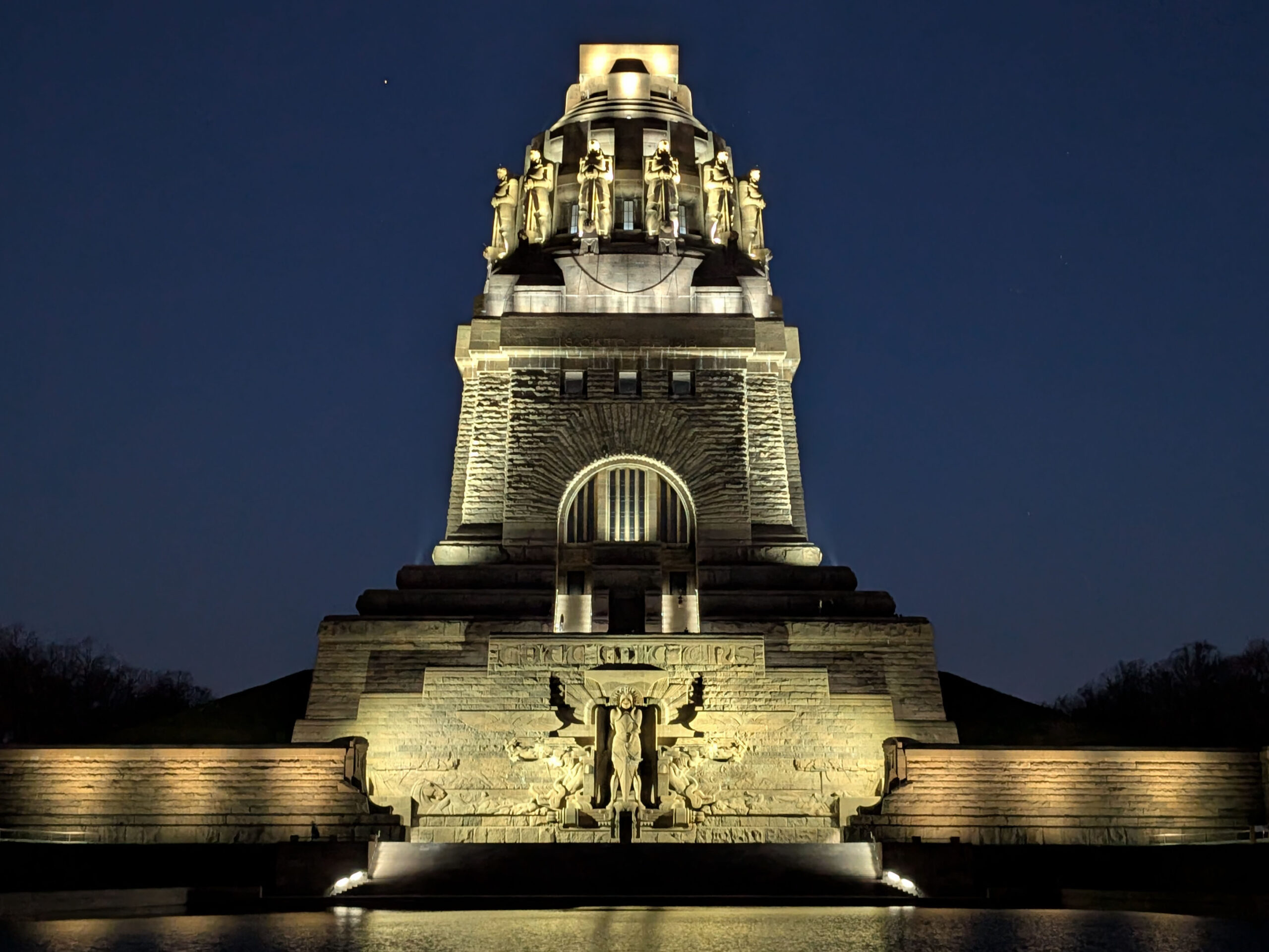 Das beleuchtete Völkerschlachtdenkmal in Leipzig, Deutschland, erhebt sich bei Nacht, und an seiner massiven Steinfassade sind Statuen und detaillierte Schnitzereien zu sehen.