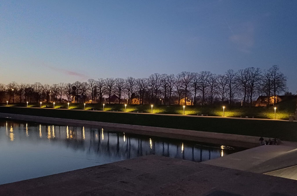 Eine heitere Abendszene zeigt eine Reihe von blattlosen Bäumen, die sich gegen den Dämmerungshimmel abheben, wobei sich die Lichter der Gehwege auf dem ruhigen Wasser eines Kanals spiegeln und ein paar Menschen am Ufer sitzen.