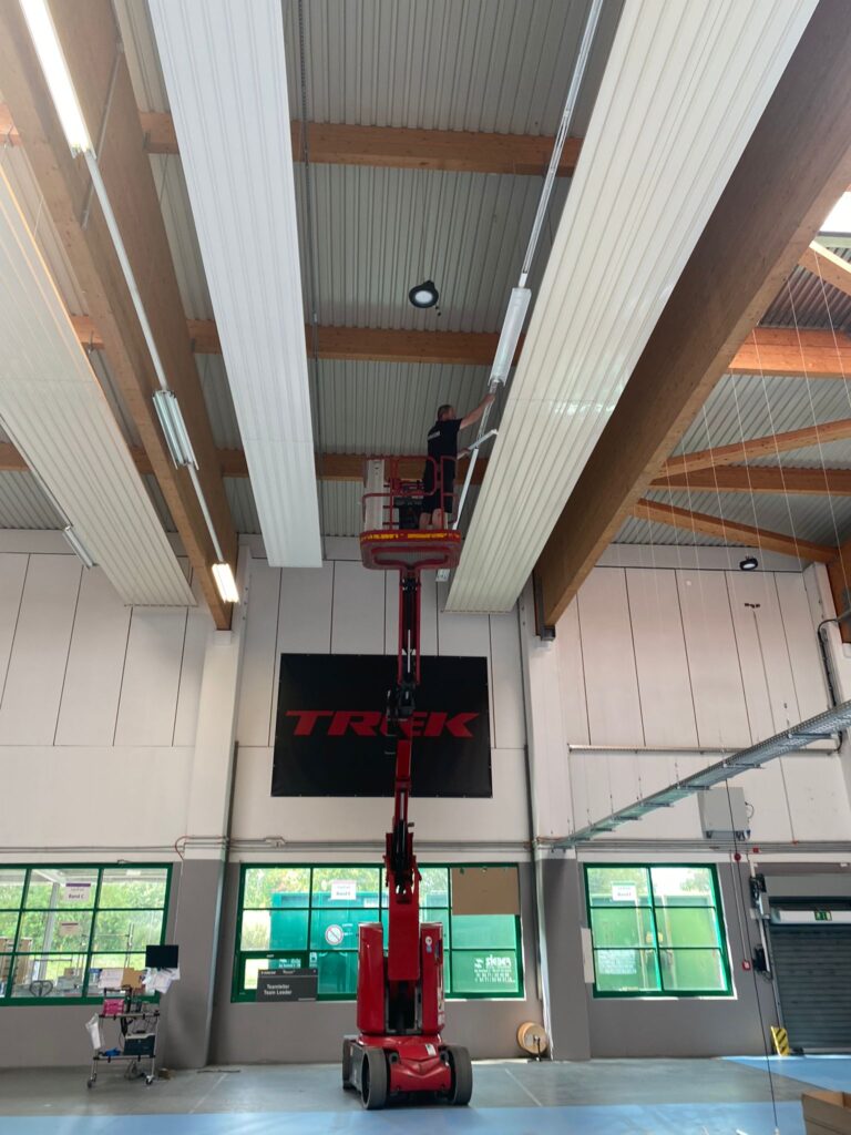 Eine Person auf einer roten Scherenbühne arbeitet nahe der Decke eines großen Innenraums mit freiliegenden Balken und Leuchtstoffröhren. An der Wand darunter sind große Fenster und ein schwarzes „TREK“-Schild zu sehen.