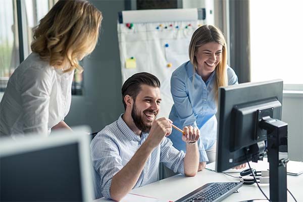 Drei Kollegen sitzen in einem Büro und arbeiten fröhlich an einem Schreibtisch mit einem Computer zusammen. Ein Mann sitzt da, hält einen Bleistift in der Hand und lächelt, während zwei Frauen daneben stehen und ebenfalls lächeln. Hinter ihnen befindet sich ein Whiteboard mit Notizen und Diagrammen zum Thema Leuchtenhersteller und innovative LED-Technik.