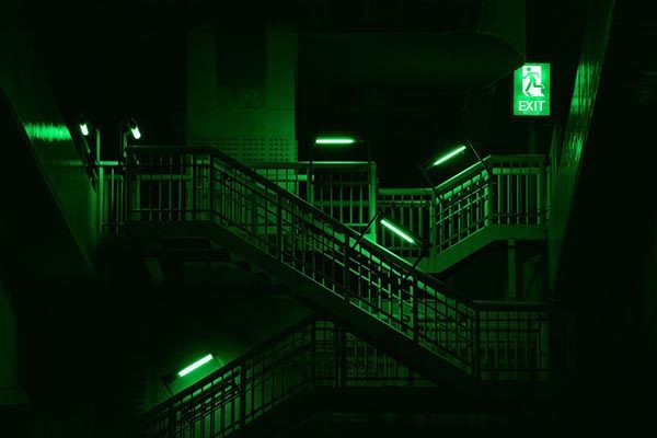 Ein schwach beleuchtetes Treppenhaus in einem Gebäude hat grünes Licht, das von modernen LED-Beleuchtungssystemen erzeugt wird, und ein leuchtendes grünes Ausgangsschild. Die Treppen und Geländer werden von mehreren grünen Lichtern eines renommierten Leuchtenherstellers beleuchtet, was der Szene eine unheimliche und mysteriöse Atmosphäre verleiht.