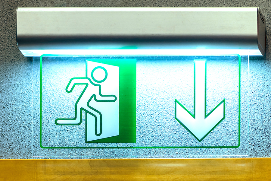 Grün beleuchtetes Notausgangsschild, das eine Person zeigt, die durch eine Tür mit einem großen, nach unten zeigenden Pfeil läuft, montiert an einer Wand über einer Holzfläche.